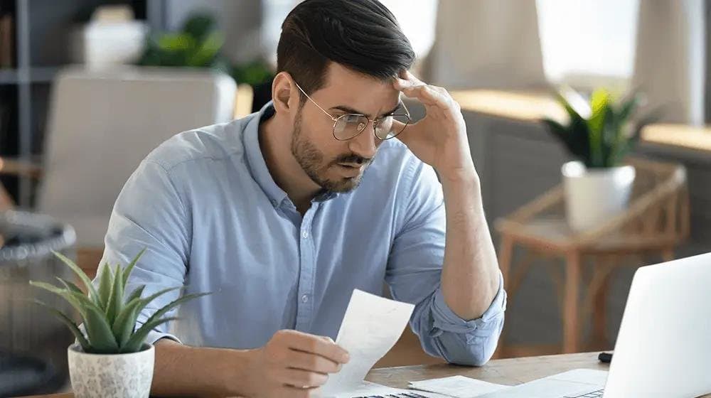 A man in glasses and a light blue shirt at his desk, rubbing his forehead as he reads a printed document beside an open laptop