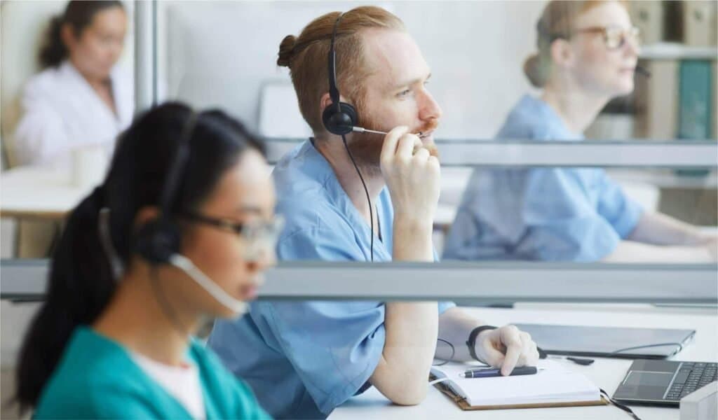 Healthcare staff in scrubs wearing headsets, taking calls at desks separated by glass partitions