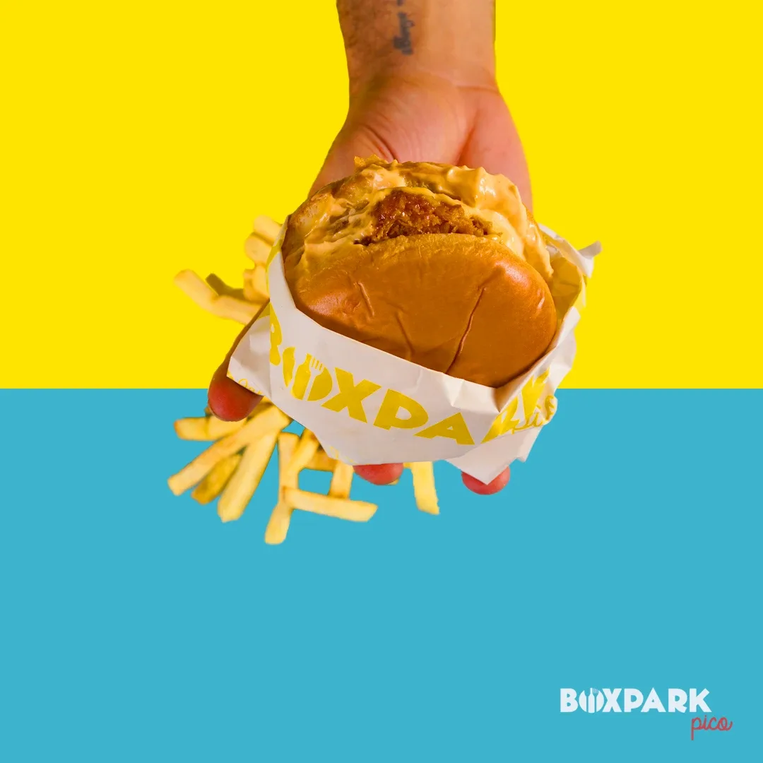 Hand holding a burger in a yellow BoxPark wrapper over yellow and blue