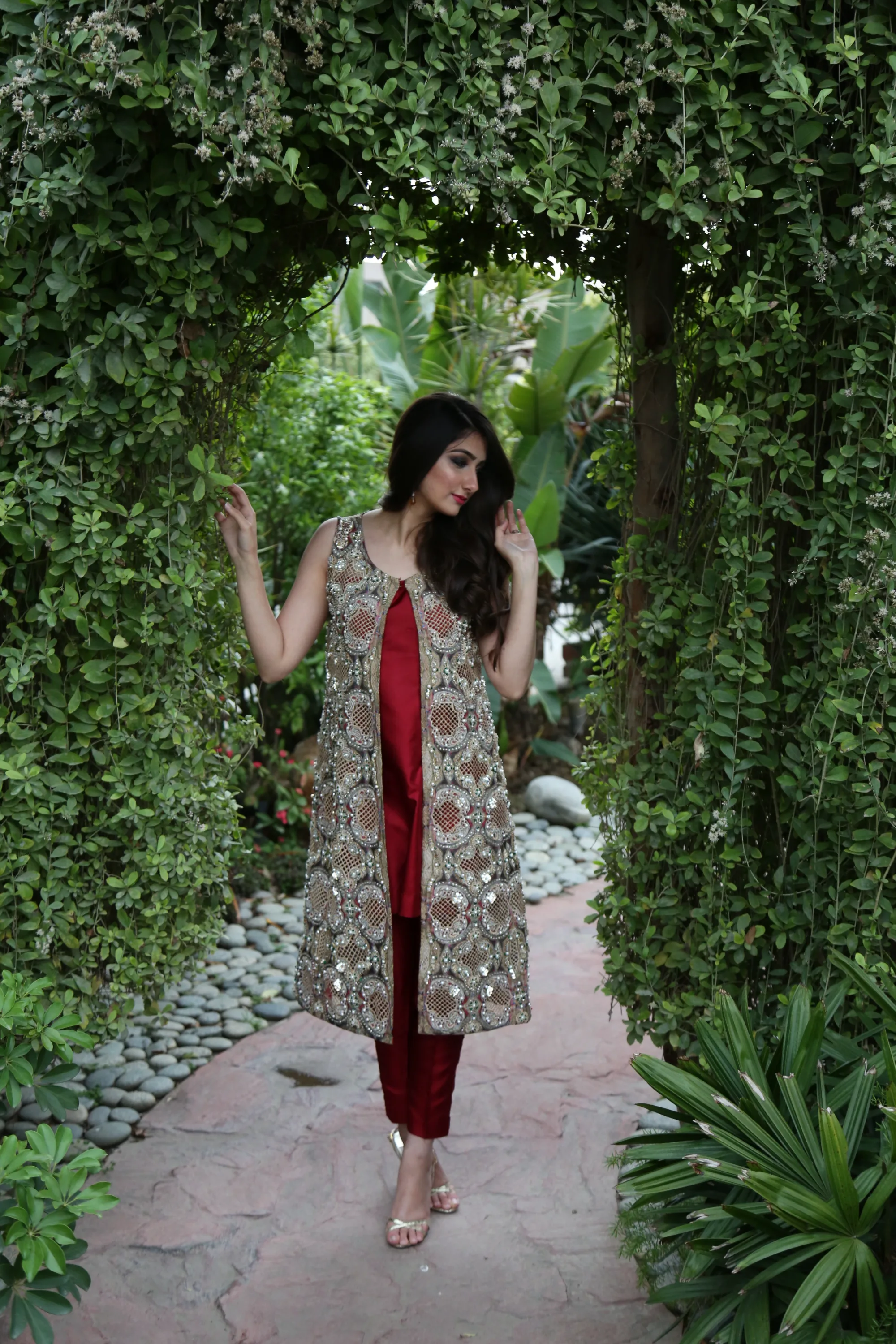 Model in a red outfit and embroidered jacket under a leafy garden arch