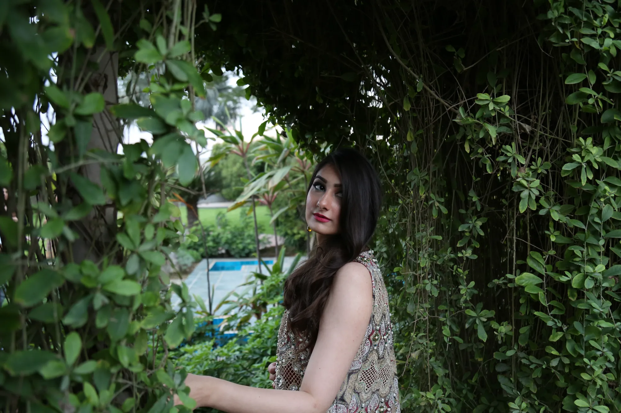 Model in embroidered jacket looking back among hanging vines in a garden